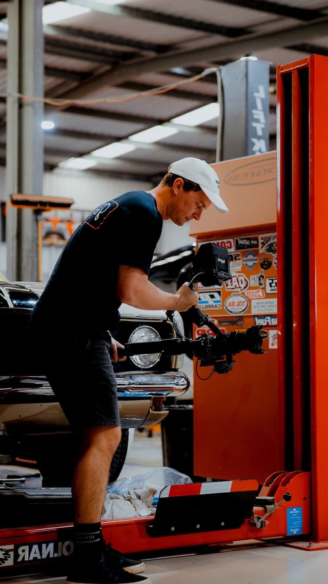 A videographer in a white cap operates a camera rig beside a classic car in a sticker covered workshop.