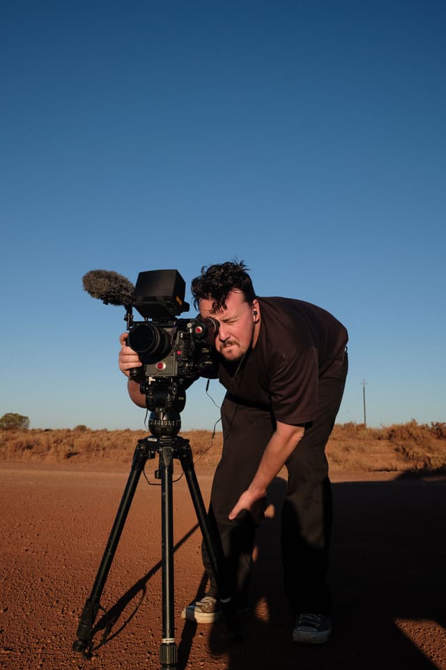 A cinematographer bends to check a red cinema camera mounted on a tripod under a clear blue outback sky.