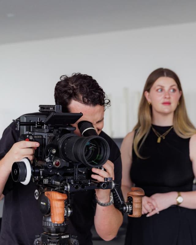 A cinematographer looks through a large cinema camera while a woman in black stands beside him in a white studio.