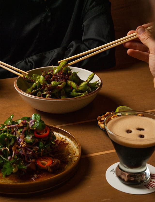 A diner uses chopsticks to eat chili edamame beside a herb salad and an espresso martini on a wooden table.