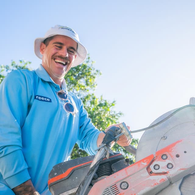A tradesperson in a Reece branded shirt and bucket hat laughs while operating a cutting saw outdoors.
