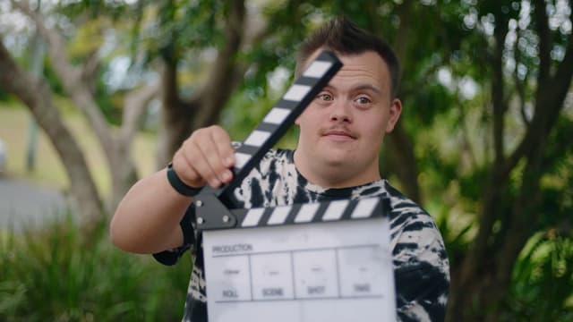 A young man holds up a clapperboard on a film set, smiling amid lush green garden foliage.
