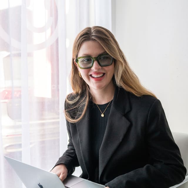 A woman in a black blazer smiles while working on a laptop near a bright window.