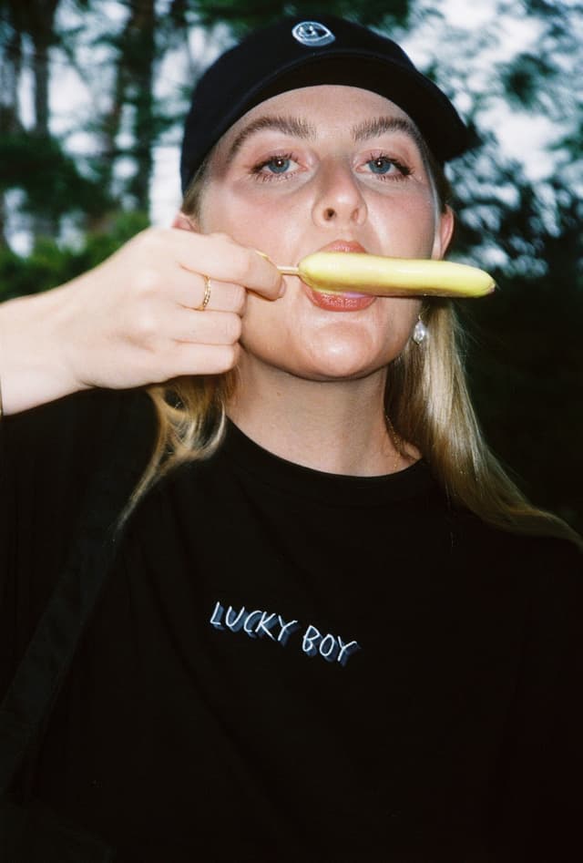 A woman in a Lucky Boy cap and t-shirt eats a yellow icy pole outdoors.