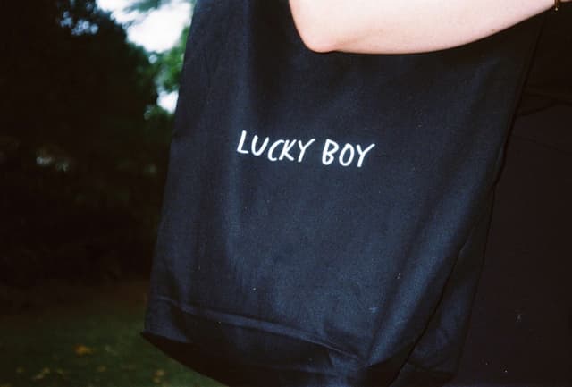 A black tote bag printed with the Lucky Boy Agency wordmark hangs from someone's shoulder outdoors.