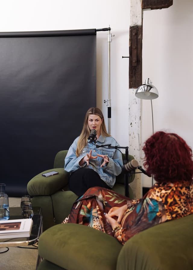 Two women sit in green armchairs recording a podcast interview with microphones on stands in a studio.