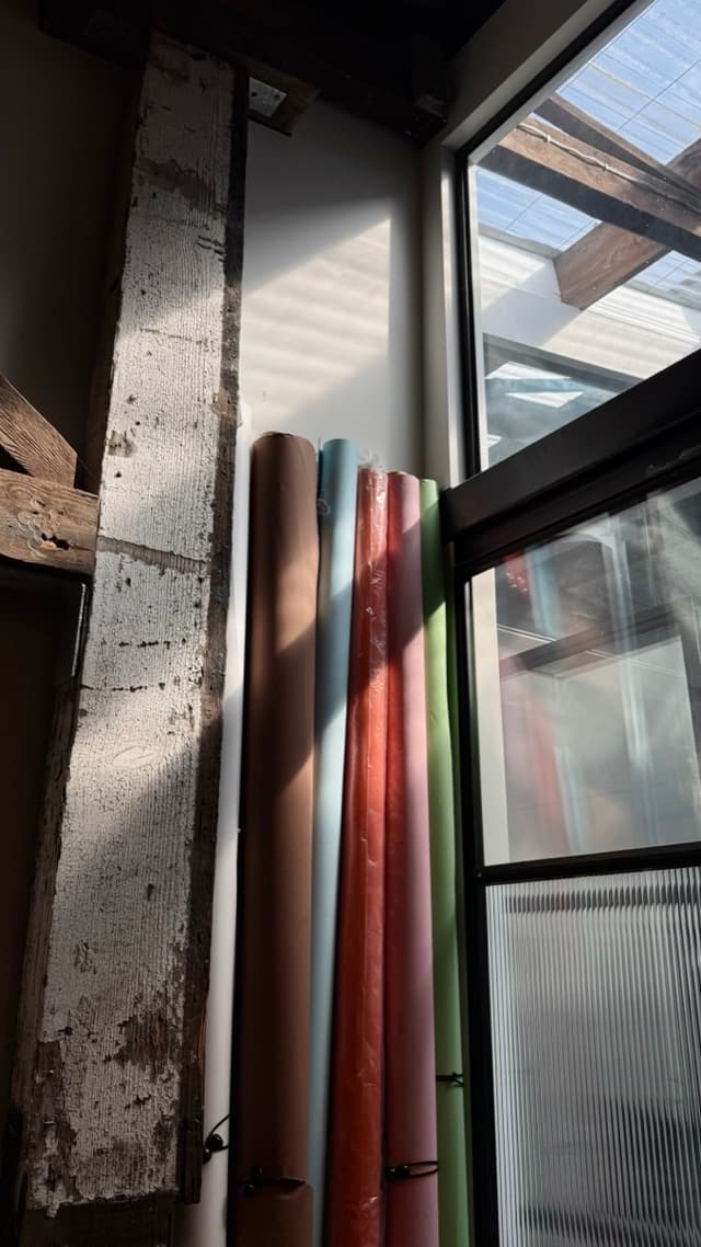 Rolls of coloured paper lean against a sunlit window beside an exposed timber beam in a design studio.