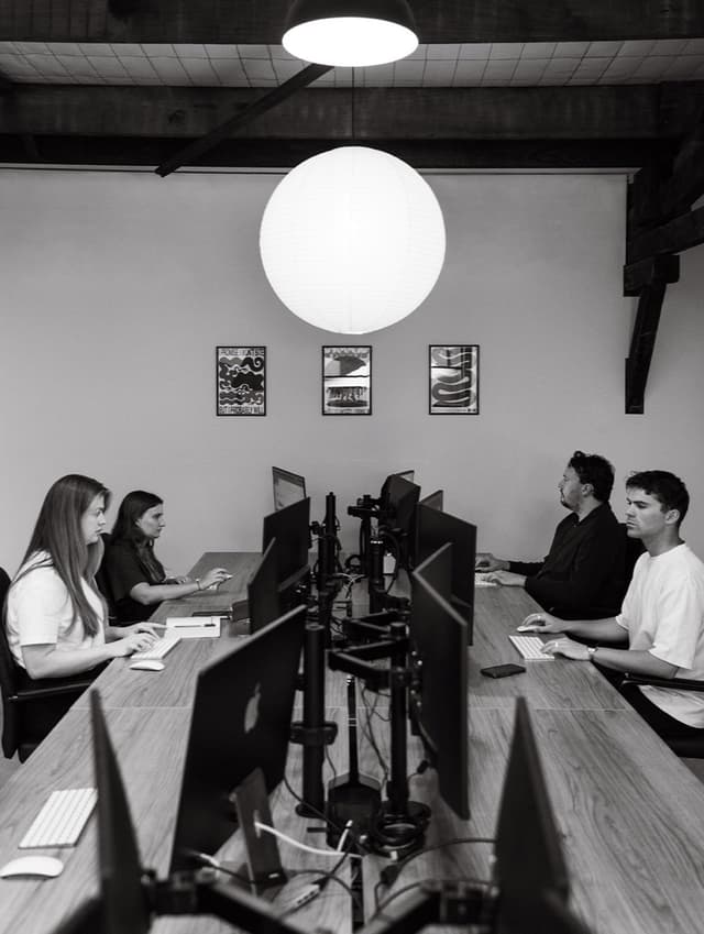 In black and white, a creative team works at multi-monitor desks in a studio lit by a round pendant lamp.