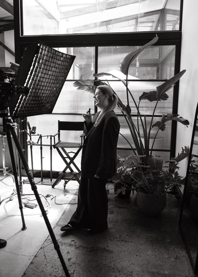 A woman laughs on set beside a camera, softbox light, and director's chair in a black and white studio photo.