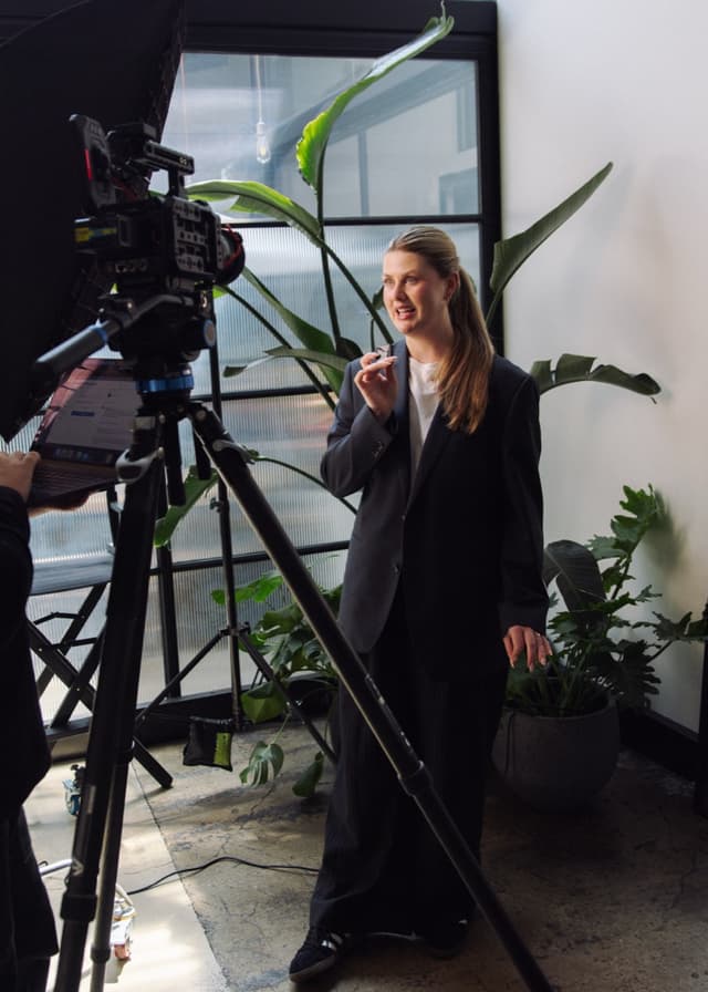 A woman speaks to camera in a studio decorated with large green plants and string lights.