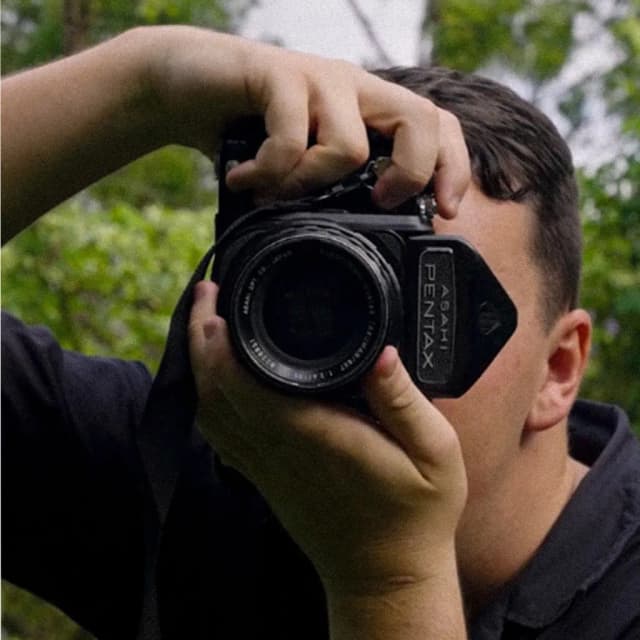 A photographer looks through a vintage Pentax camera framed by green trees behind him.