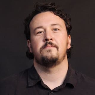 A studio headshot shows a man with curly dark hair and a beard against a plain dark background.