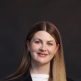 A professional headshot of a woman with long brown hair wearing a black blazer over a white top.
