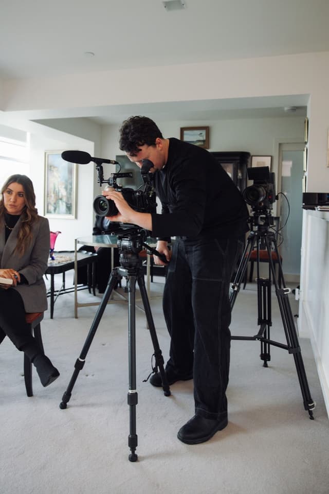 A camera operator films a seated woman being interviewed in a home lounge room with two tripod-mounted cameras.