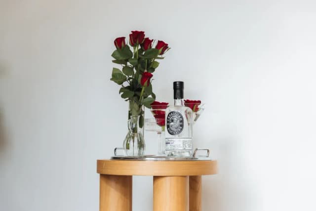 A bottle of Capricorn Distilling Co spirit stands beside a vase of red roses and a martini glass on a side table.