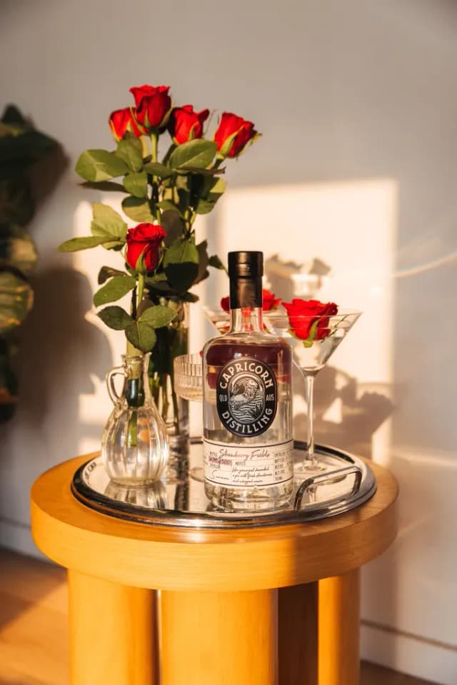 A Capricorn Distilling Co Strawberry Fields gin bottle with red roses in vases on a round side table.