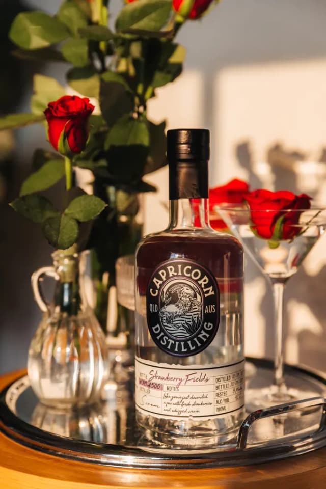 A bottle of Capricorn Distilling Co Strawberry Fields gin sits beside red roses and a martini glass.
