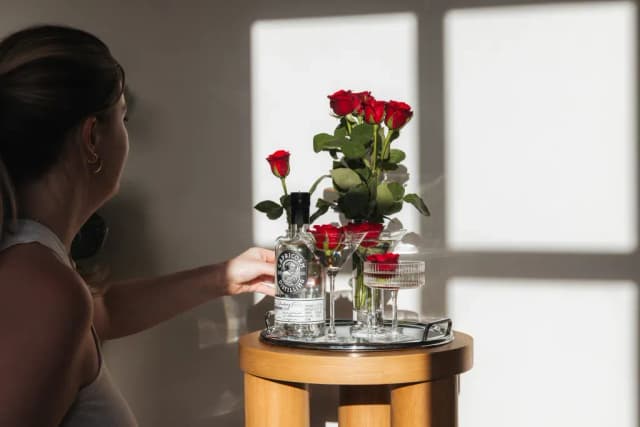 A woman reaches for a Capricorn Distilling gin bottle staged with roses on a side table.