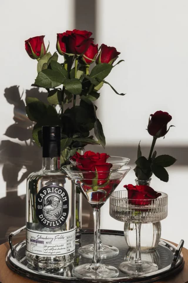 A Capricorn Distilling Co Strawberry Fields gin bottle beside red roses in glasses and vases on a tray.