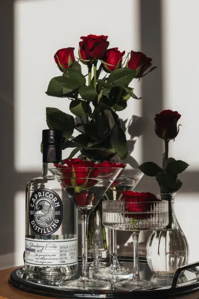 A Capricorn Distilling Co gin bottle surrounded by red roses and cocktail glasses on a bar tray.