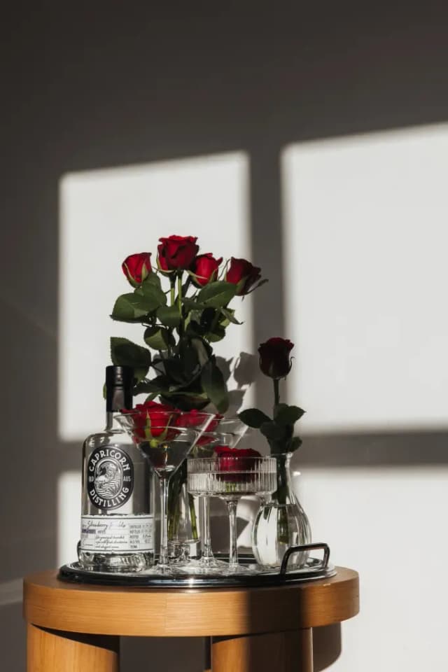 A Capricorn Distilling gin bottle staged with red roses and martini glasses in warm window light.