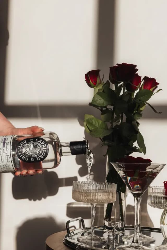 A hand pours Capricorn Distilling Co gin into glasses beside a bouquet of red roses in sunlight.