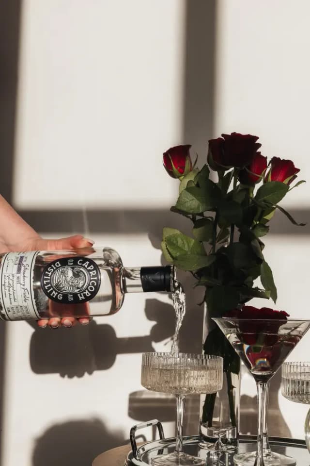 A hand pours Capricorn Distilling Co spirit into glasses beside red roses, sunlight casting shadows across the table.