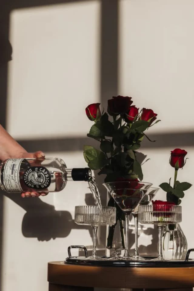 A hand pours Capricorn Distilling Co spirit into martini glasses beside a bouquet of red roses in sunlight.