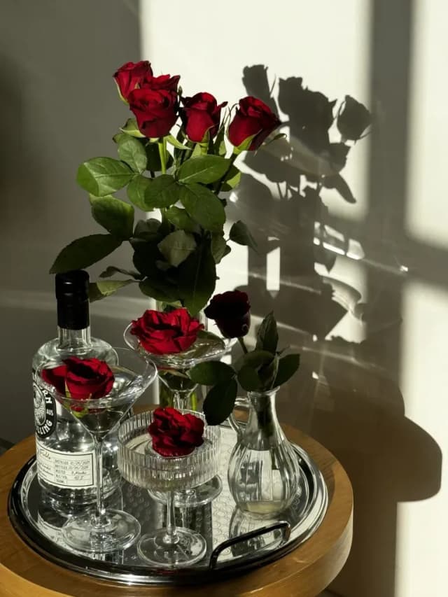 A Capricorn Distilling gin bottle and martini glasses surrounded by red roses on a sunlit tray.