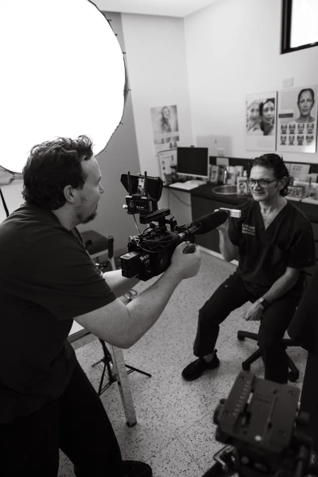 Camera operator filming a seated subject during a talking-head interview with a softbox lighting the room.