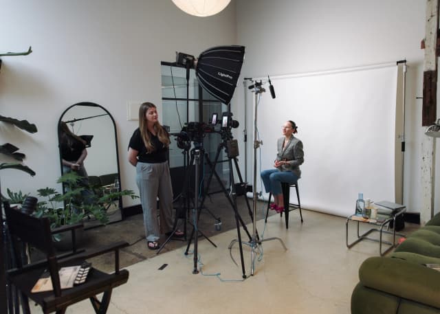 A crew member operates a camera rig beside a softbox light while a woman is interviewed on a stool in a studio.