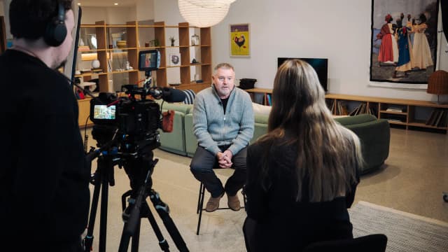 A film crew records an interview with a seated man in a living room styled with green sofas and artwork.