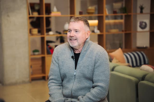A man in a grey zip up sweater speaks during an on camera interview in front of a bookshelf and sofa.