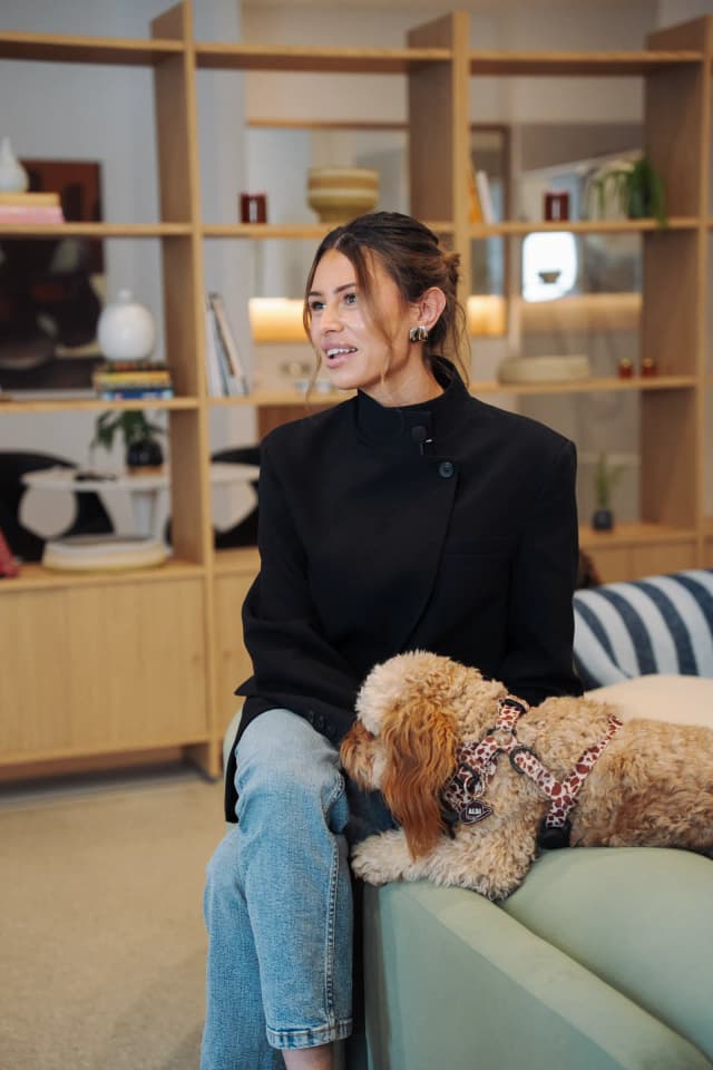 A woman in a black blazer sits smiling on a couch as a golden doodle rests against her leg.
