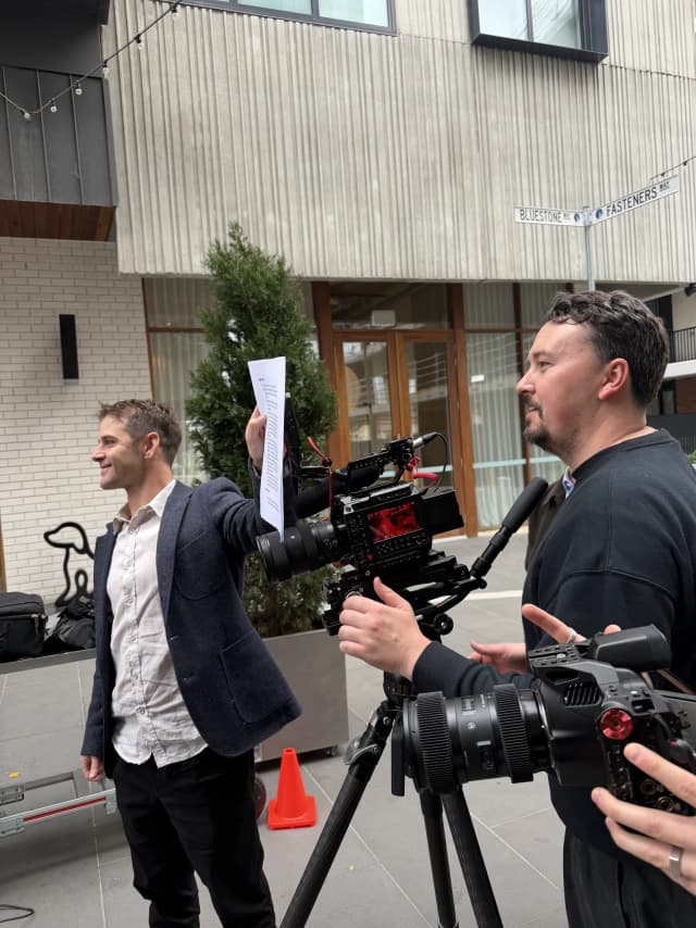 Two crew members review a shot list beside a cinema camera on a tripod, with Bluestone Way and Fasteners Way street signs behind them.