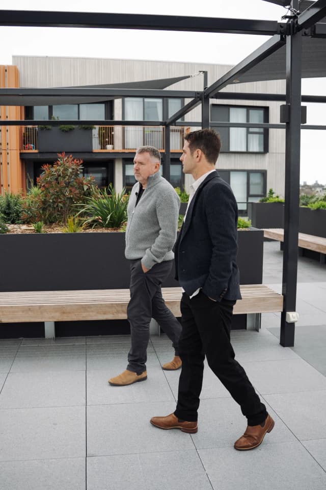 Two men walk and talk across a rooftop terrace lined with garden beds and apartment buildings.