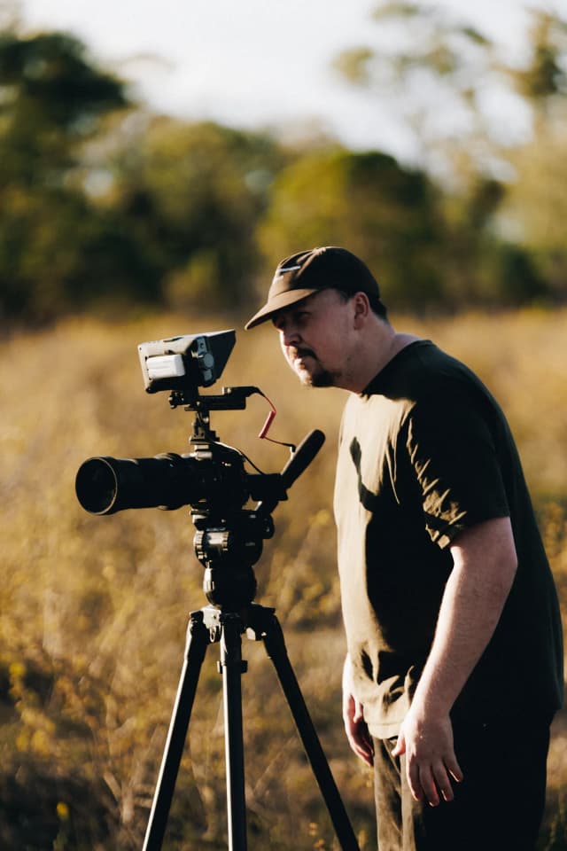 A videographer looks through a camera on a tripod in a golden grass field, from a Providore Global farm shoot.