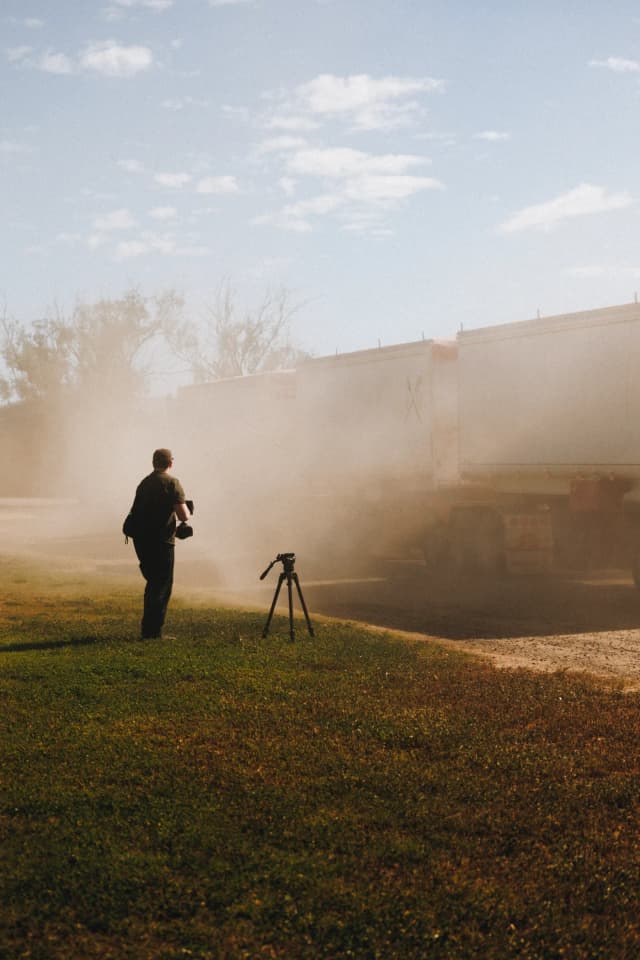 A camera operator with a tripod films a dusty truck on a Providore Global farm at golden hour.