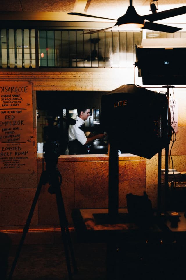 A chef cooks in a restaurant kitchen while film lighting and a tripod are set up nearby, a Providore Global shoot.