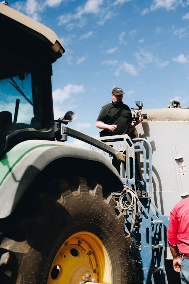 A cameraman films from a tractor's platform beside a man in a red shirt on a Providore Global farm.
