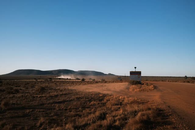 A dusty outback road for Australia Post stretches toward low hills, with a signpost and dust cloud in the distance.