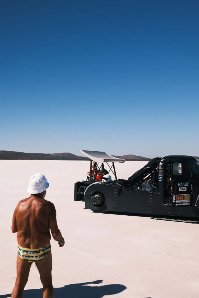 A man in a bucket hat and swimwear walks across the salt flat toward a black streamlined race car.