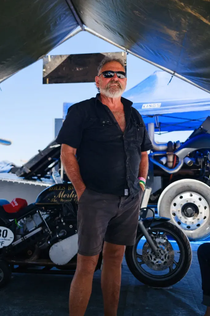A man in sunglasses stands beside a vintage drag racing motorcycle under a marquee at a salt lake speed event.