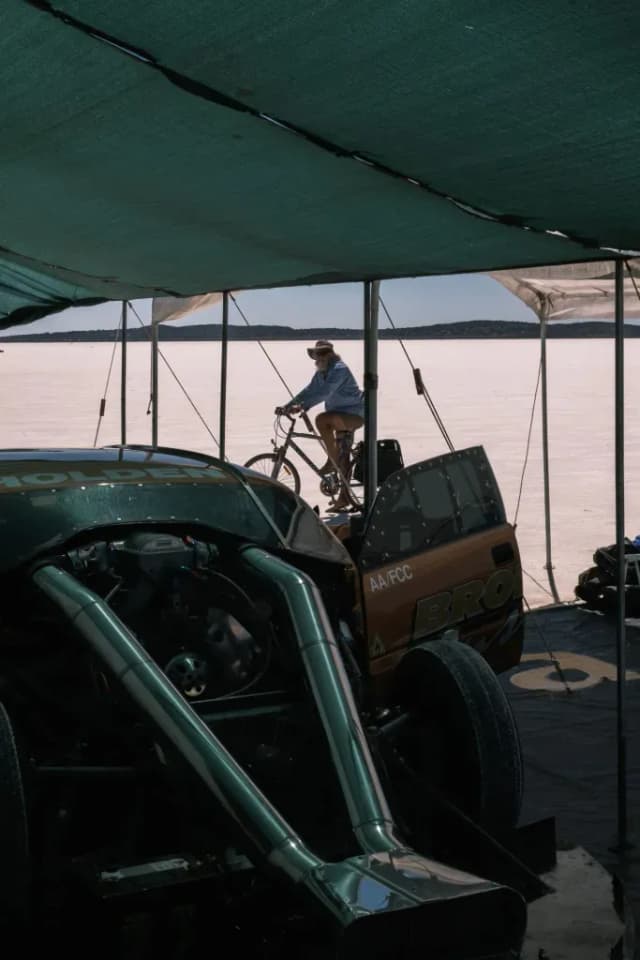 A person rides a bicycle beside a vintage green drag racer parked under a canopy on a dry lake bed.