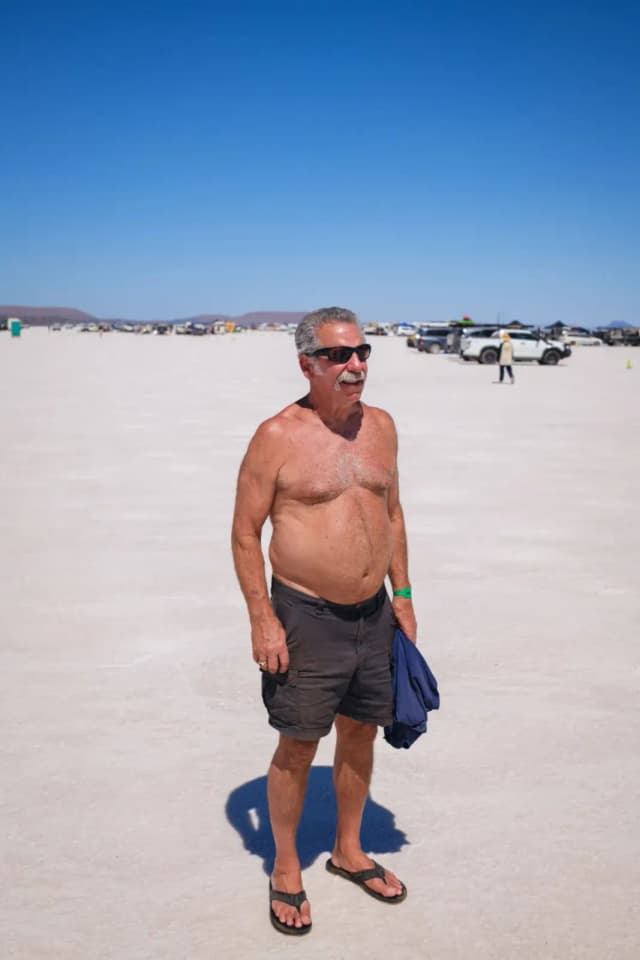 A shirtless man in sunglasses and shorts stands on the vast white salt flat with vehicles parked in the distance.
