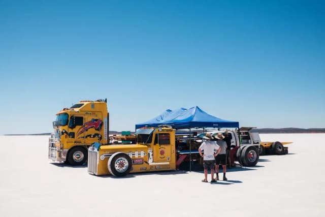 Two men stand beside vintage yellow Kenworth trucks and a blue marquee on the white salt lake.