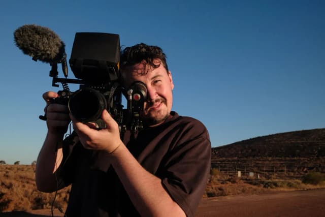 A cameraman holds a RED cinema camera to his eye, smiling, with a dry outback landscape behind him.