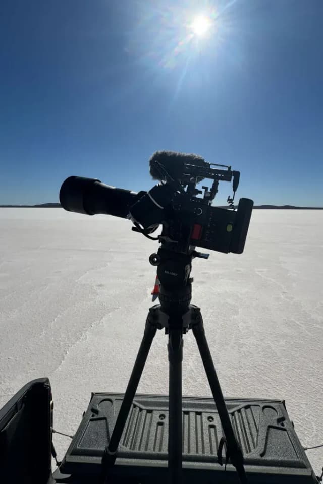 A cinema camera on a tripod faces a vast white salt flat under a bright sun.