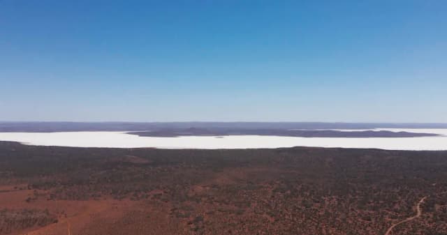 An aerial view of a white salt lake surrounded by red outback scrubland under a clear blue sky.