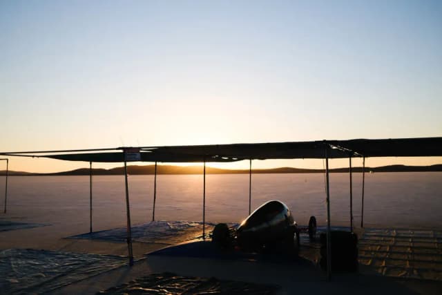 A streamlined land speed racing car sits under a canopy on the salt flats at sunset.